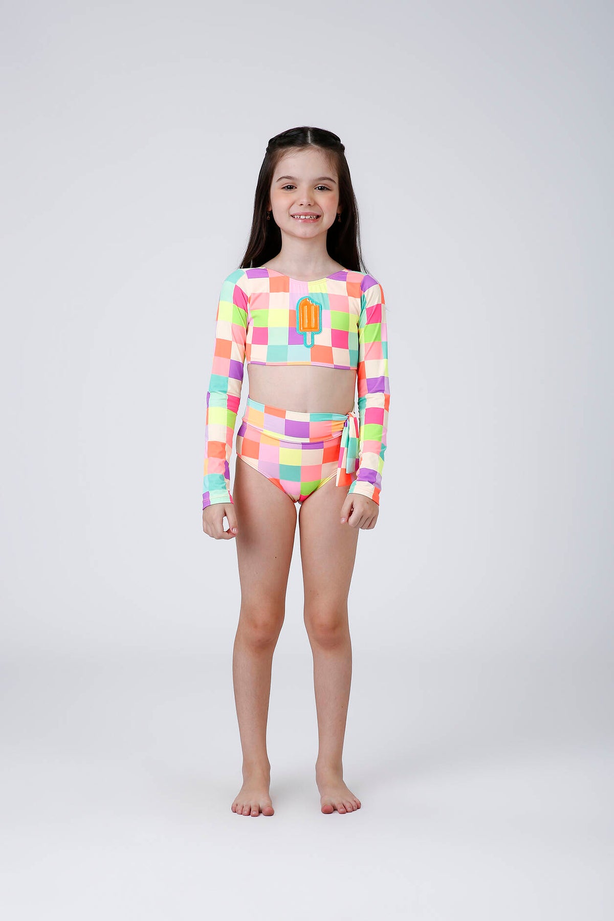 Menina usando biquíni infantil Thais Picolé Siri com xadrez multicolorido, blusa de manga longa com aplique de picolé e calcinha de cintura alta com laço lateral.