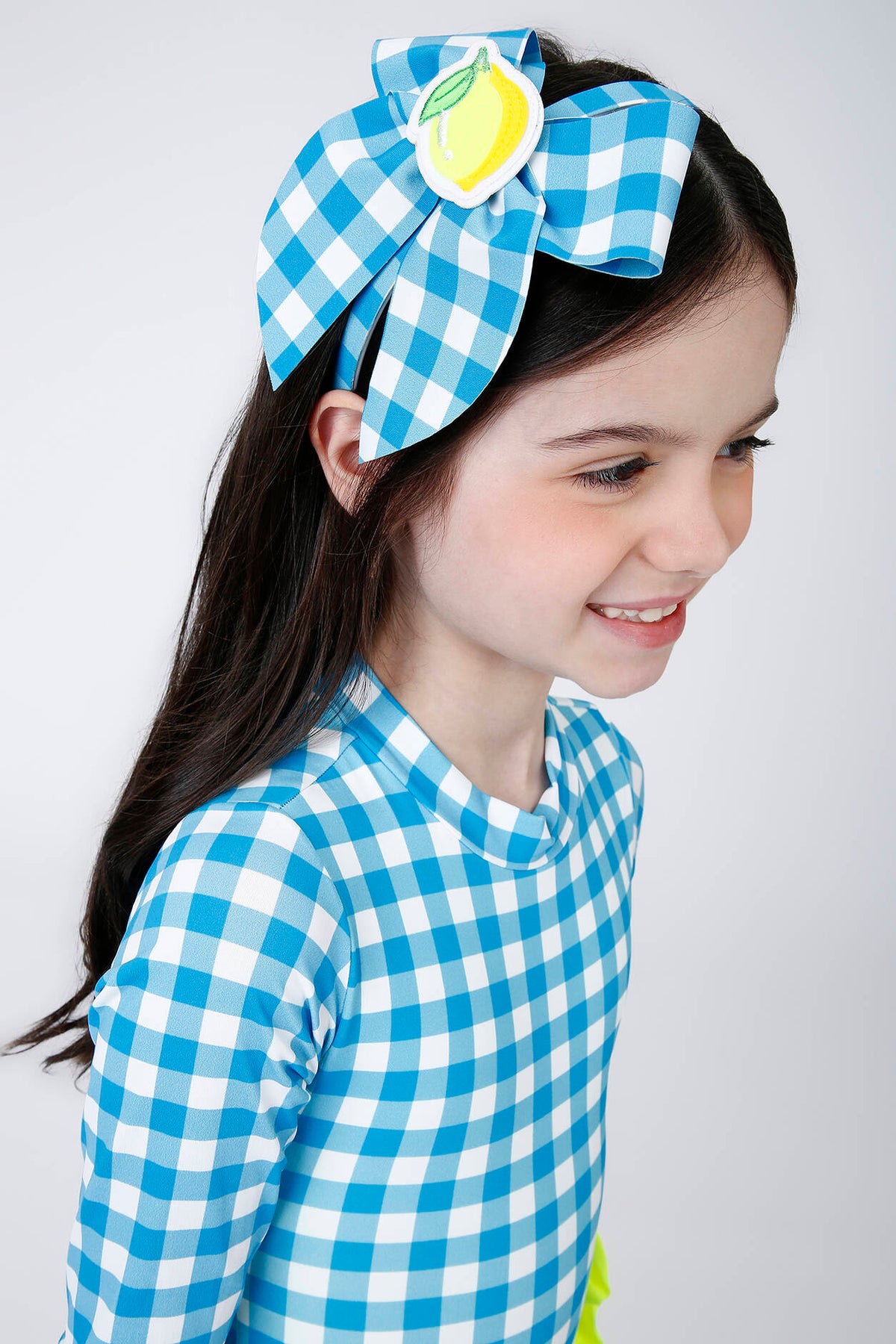 Menina usando tiara infantil Siri com laço xadrez azul e branco e aplique de limão amarelo com folha verde.
