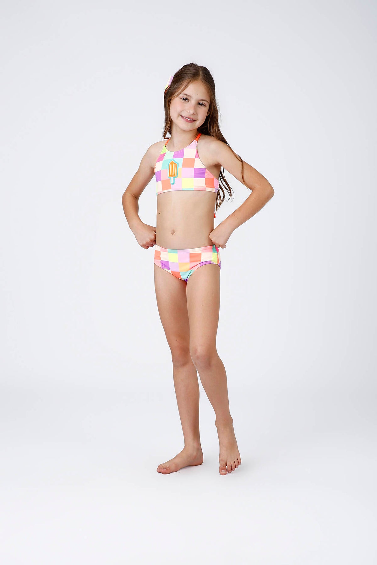 Menina usando biquíni infantil Tamires Picolé Siri com xadrez multicolorido, top com aplique de picolé e calcinha na mesma estampa.